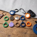 3D printed AirTag holders in green, lilac, tan, navy, and orange — keychain and pet collar versions with engraved name plaque