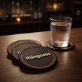 Four matte black 3D-printed coasters fanned on a bar counter, each with a coral ring and the thingness wordmark, a cold glass resting on a fifth