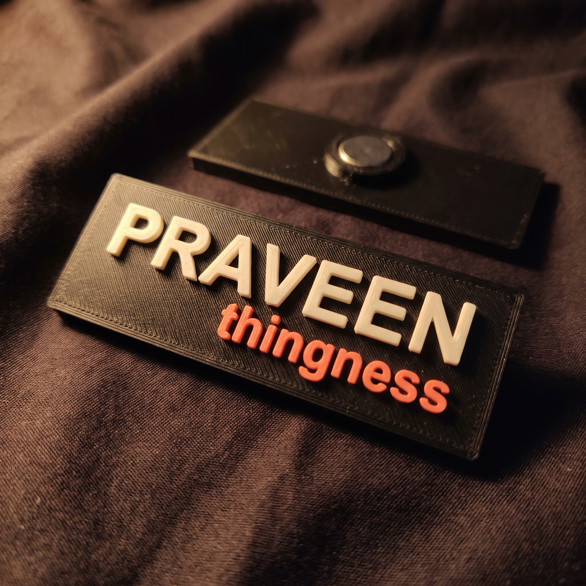 A matte black 3D-printed staff name badge on dark apron fabric, a name in raised bone lettering above the thingness wordmark in coral, with a second badge behind showing its magnetic back