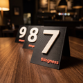 Three matte black 3D-printed table numbers reading seven, eight and nine in a row on a dark restaurant table, each with the thingness wordmark in coral on the base