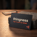 A matte black 3D-printed NFC business card propped against a stone block, the thingness wordmark in bone lettering above tap to save my details in coral, with an NFC symbol in the corner