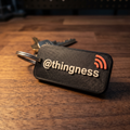A matte black 3D-printed NFC keychain tag on a dark walnut surface, raised bone-white lettering reading at thingness with a coral NFC symbol, on a steel keyring with keys behind