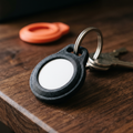 A matte black 3D-printed AirTag holder on a dark walnut surface holding a white tracker disc, on a steel keyring with keys behind and a coral holder out of focus