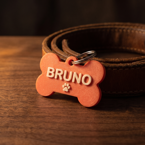 A coral bone-shaped 3D-printed pet tag reading BRUNO with a paw print, attached to a tan leather collar on a dark walnut surface