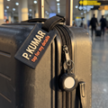The Bag Set on a suitcase — a matte black luggage tag reading P. KUMAR above tap for my details, and a matching matte black AirTag holder clipped to the zip pull in the same finish