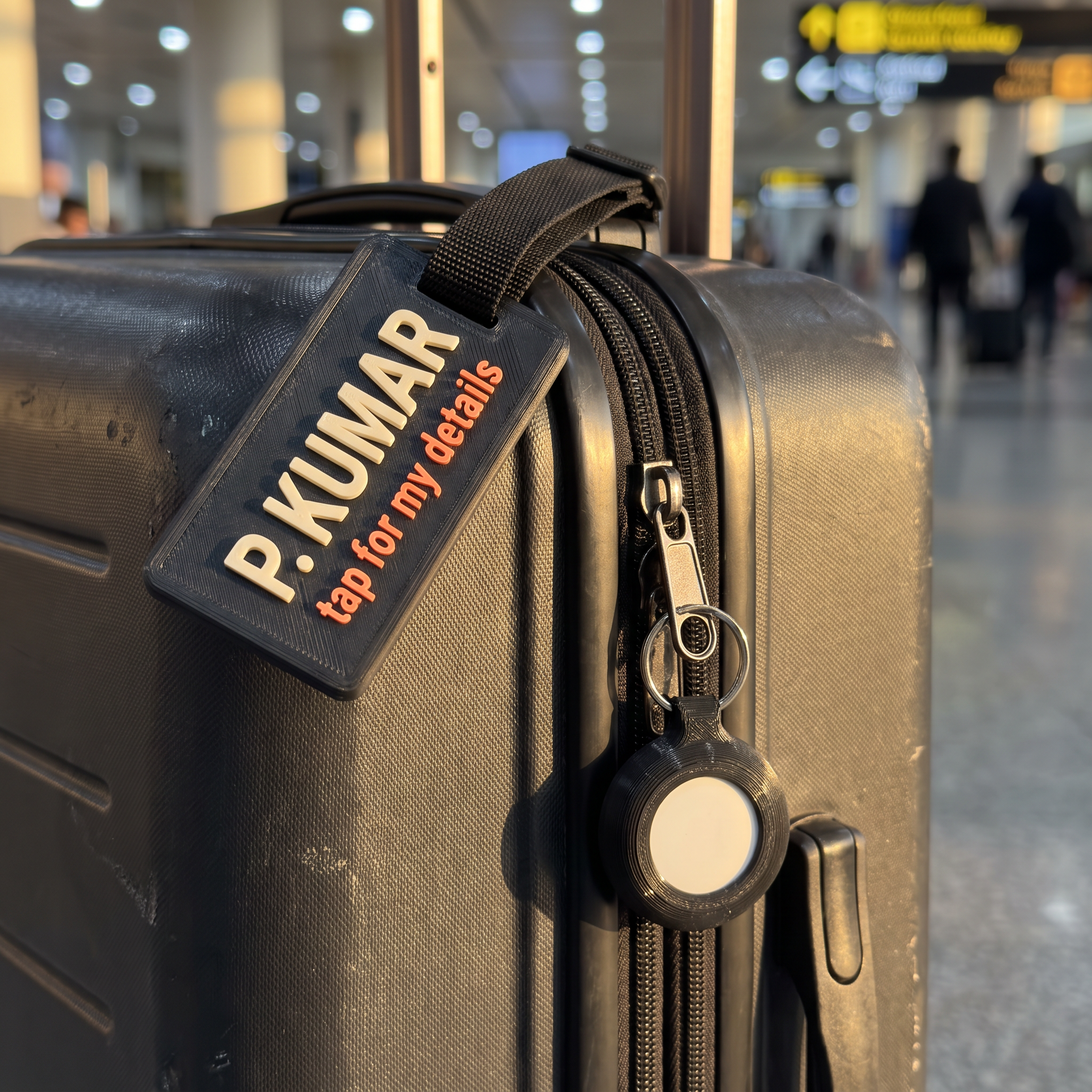 The Bag Set on a suitcase — a matte black luggage tag reading P. KUMAR above tap for my details, and a matching matte black AirTag holder clipped to the zip pull in the same finish